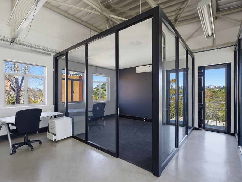 Modern office space featuring a glass-enclosed meeting room, exposed ceiling trusses, polished concrete floors, and large windows providing natural light and exterior views.