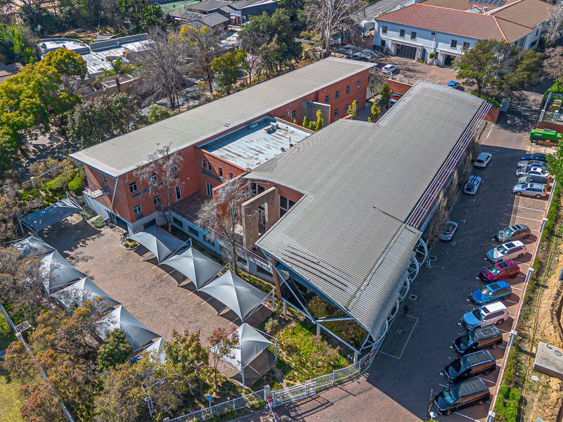 Aerial view of a commercial office complex featuring a brick building, extensive paved parking with shade sails, and surrounding landscaped grounds.