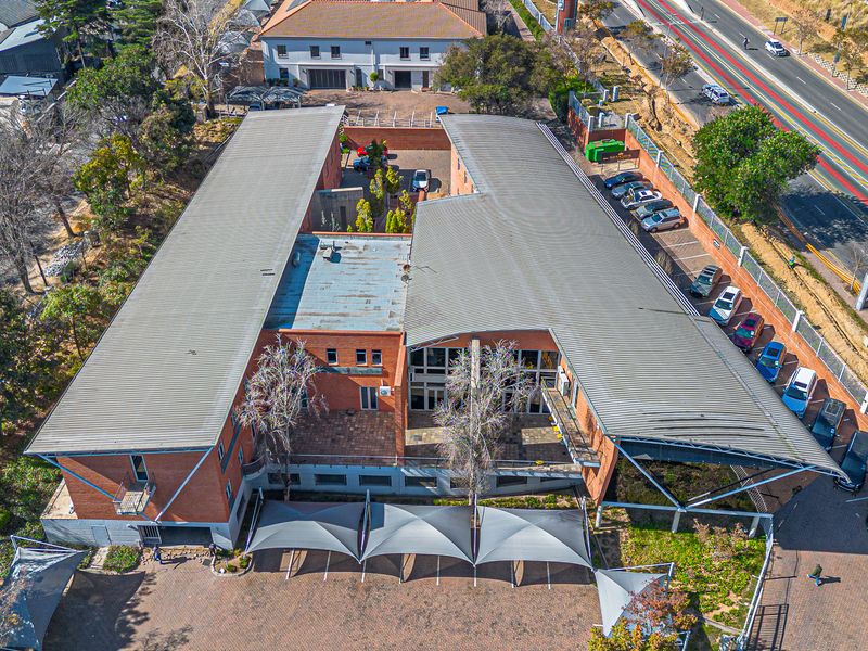 Aerial view of a commercial office building featuring a large metal roof, paved parking area with shade sails, and established surrounding landscaping.