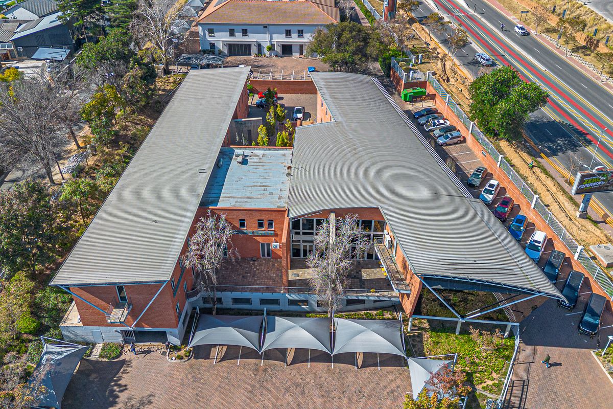 Aerial view of a commercial office building featuring a large metal roof, paved parking area with shade sails, and established surrounding landscaping.