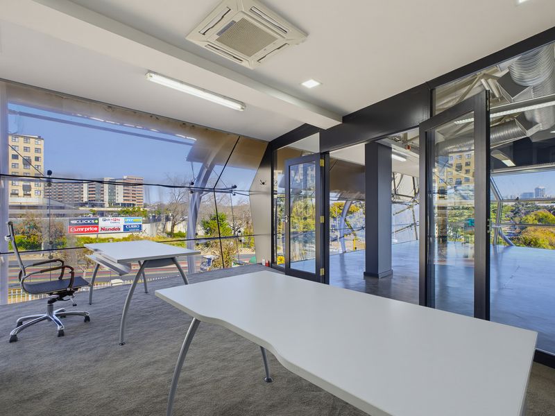 Modern office space featuring floor-to-ceiling glass walls, exposed ceiling ducting, recessed air conditioning, and carpeted flooring with views of the surrounding urban area.