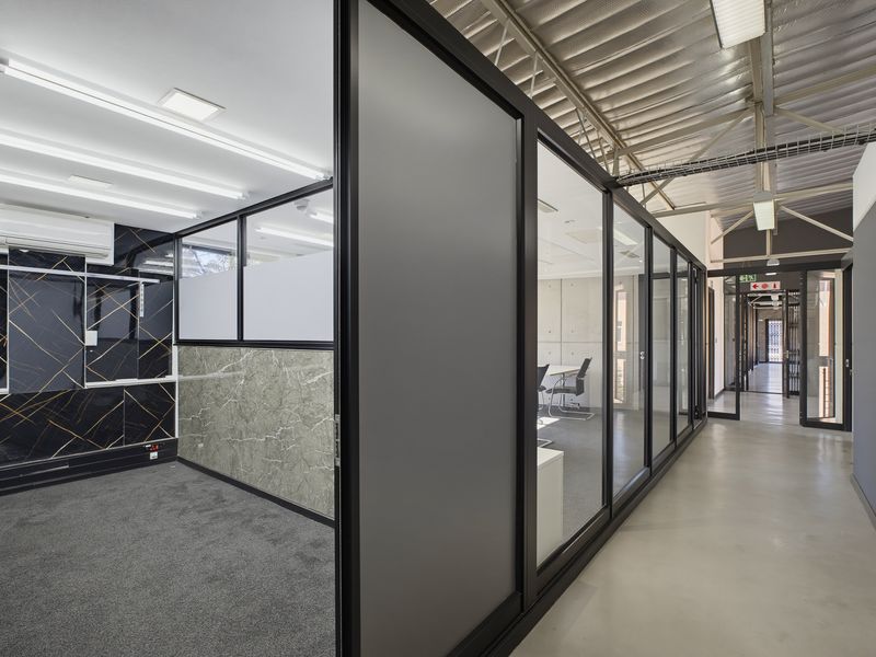 Modern office space featuring glass partitioning, exposed industrial ceilings, and a mix of carpeted and polished concrete flooring with contemporary wall finishes.