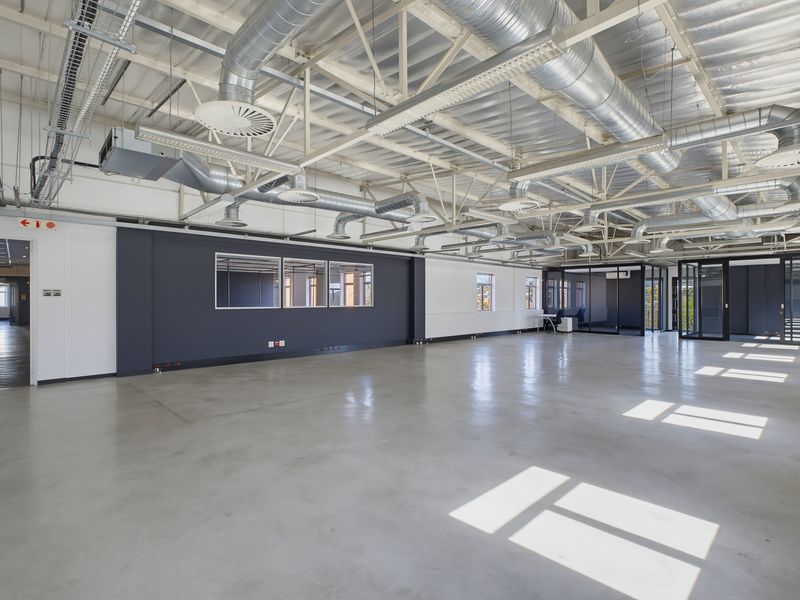 Spacious open-plan office area featuring polished concrete floors, exposed industrial-style ceiling with HVAC ducting, and modern glass partitions.