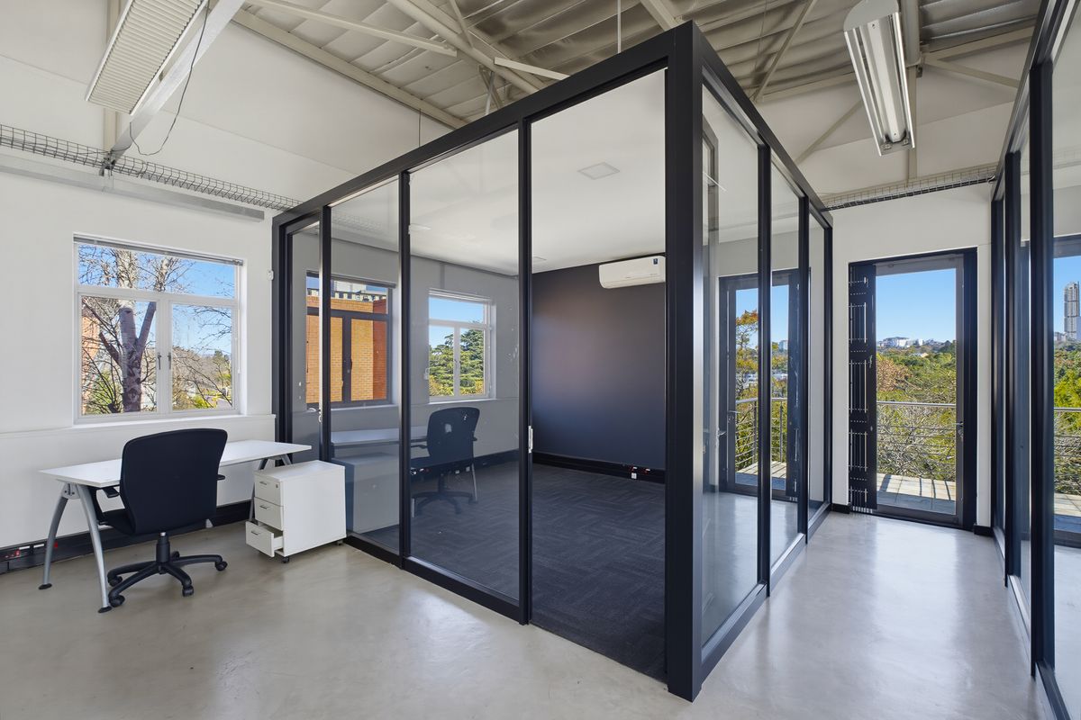 Modern office space featuring a glass-enclosed meeting room, exposed ceiling trusses, polished concrete floors, and large windows providing natural light and exterior views.