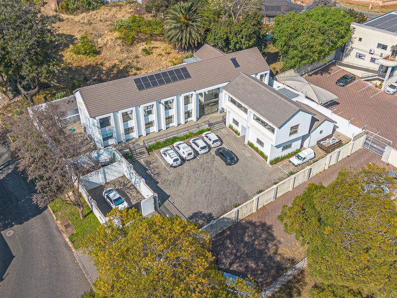 Aerial view of a commercial property featuring a multi-level building, paved parking area, solar panels, and secure perimeter fencing.