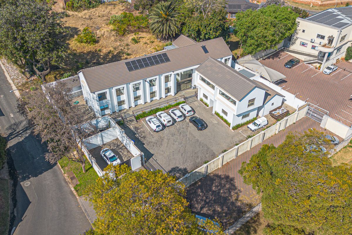 Aerial view of a commercial property featuring a multi-level building, paved parking area, solar panels, and secure perimeter fencing.