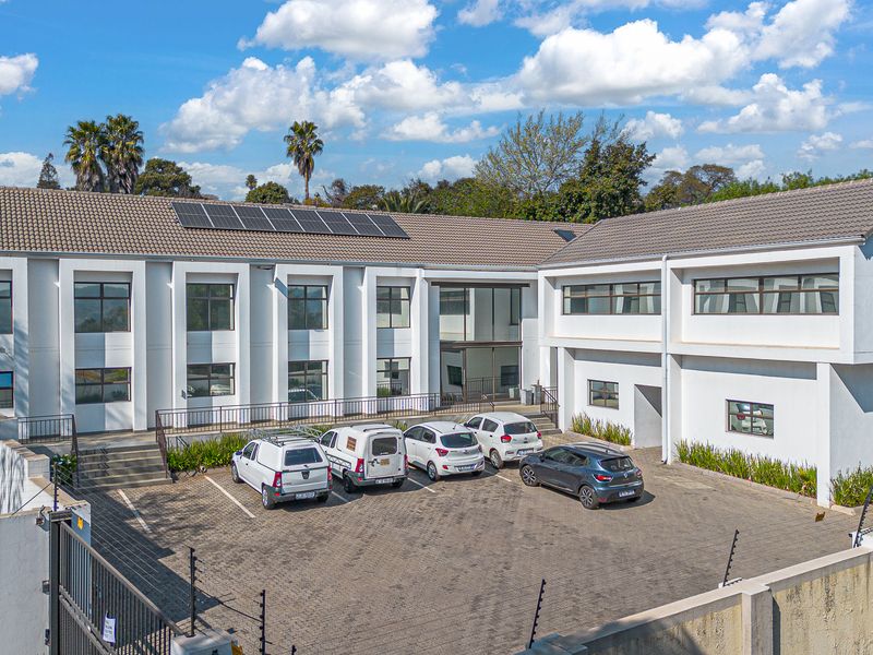 A professional aerial view of a modern commercial office building featuring a paved parking area, solar panels, and secure perimeter fencing.