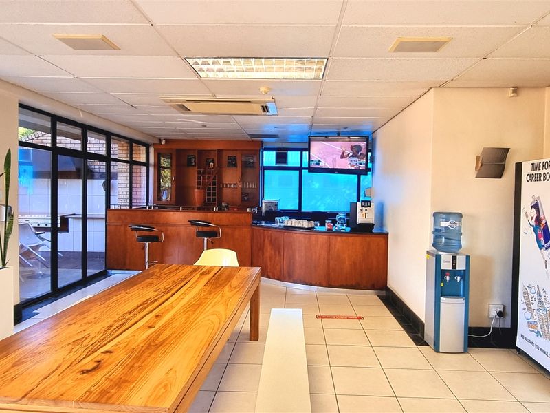 A communal office break area featuring a built-in wooden bar counter, tiled flooring, and large windows providing natural light.