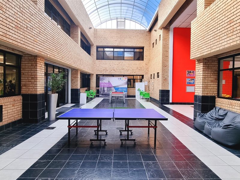 Spacious indoor atrium with a high glass vaulted ceiling, exposed brick walls, and tiled flooring, serving as a communal breakout area for the office.