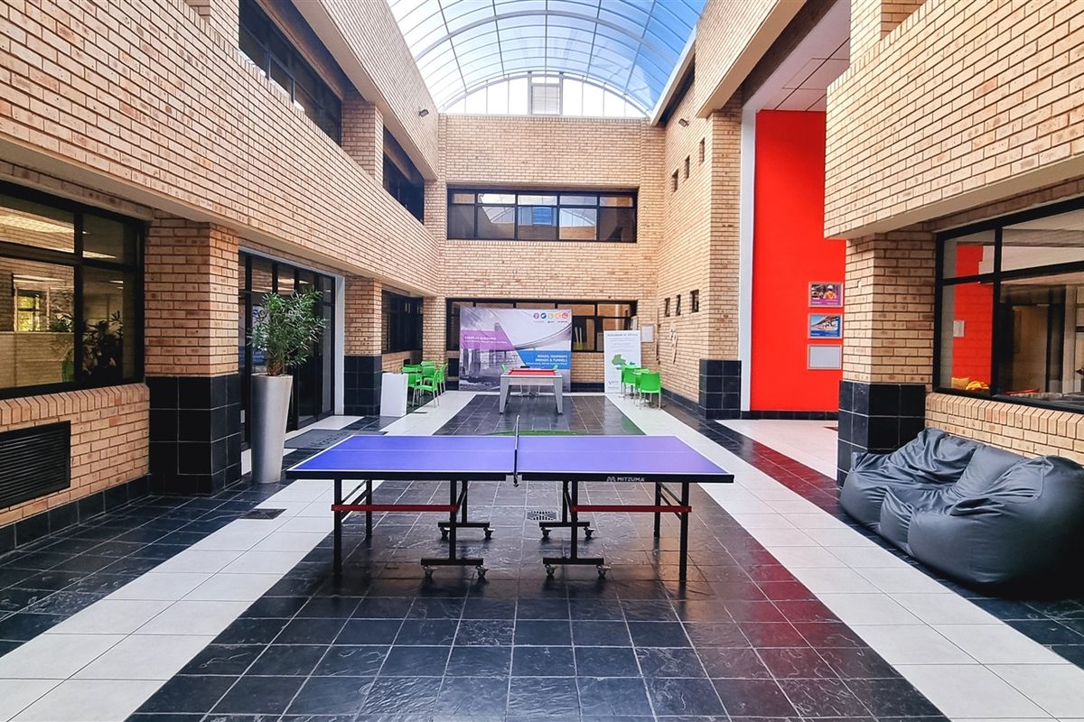 Spacious indoor atrium with a high glass vaulted ceiling, exposed brick walls, and tiled flooring, serving as a communal breakout area for the office.