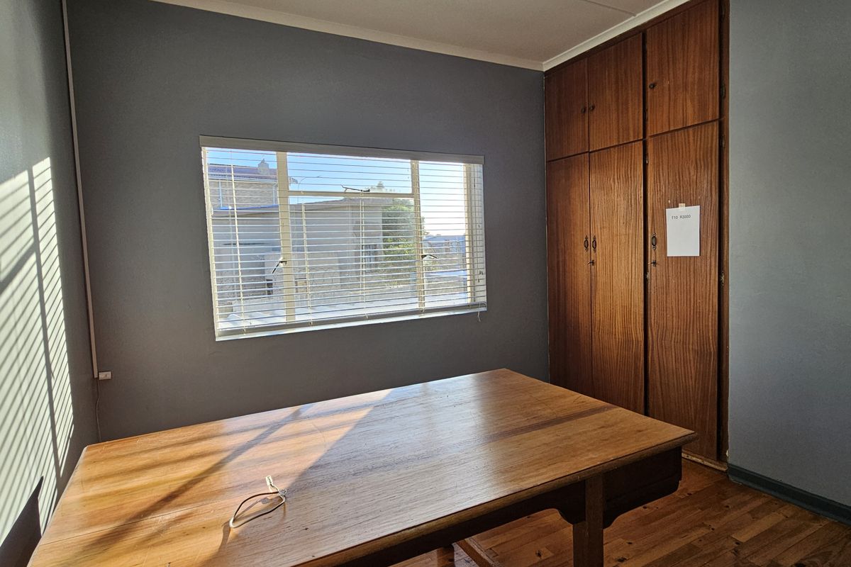 A bedroom featuring wooden flooring, built-in wooden cupboards, and a window with fitted blinds, offering a functional space with natural light.