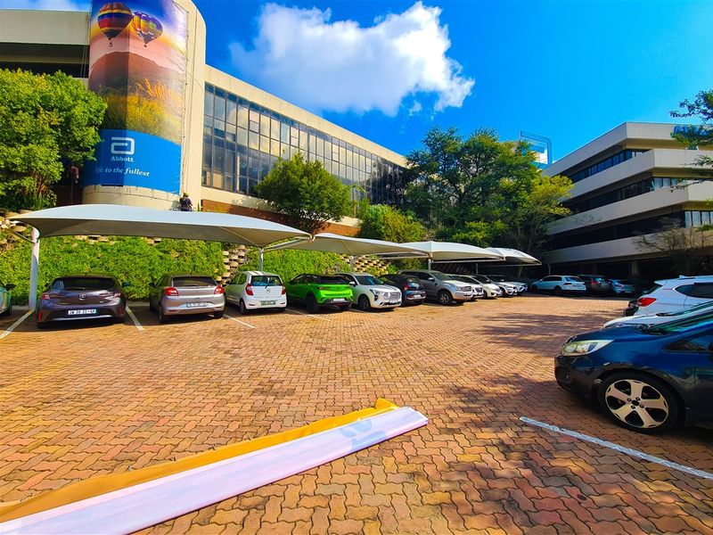 Exterior view of a commercial office building featuring a paved parking lot with shade cloth carports and landscaped retaining walls.
