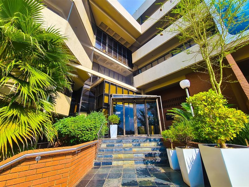 Modern multi-story office building entrance featuring a glass facade, slate tiled steps, and a brick retaining wall with integrated landscaping.