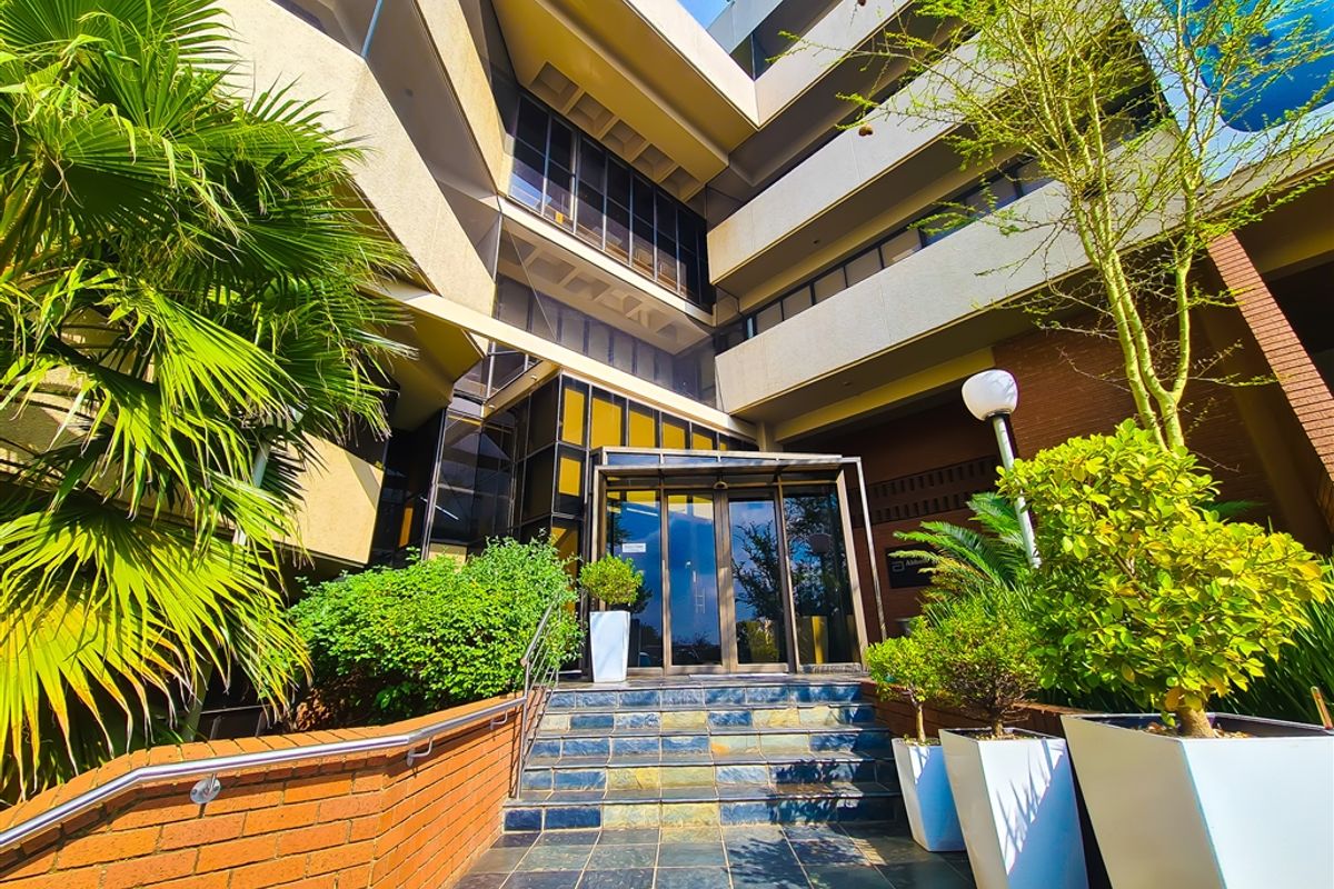Modern multi-story office building entrance featuring a glass facade, slate tiled steps, and a brick retaining wall with integrated landscaping.