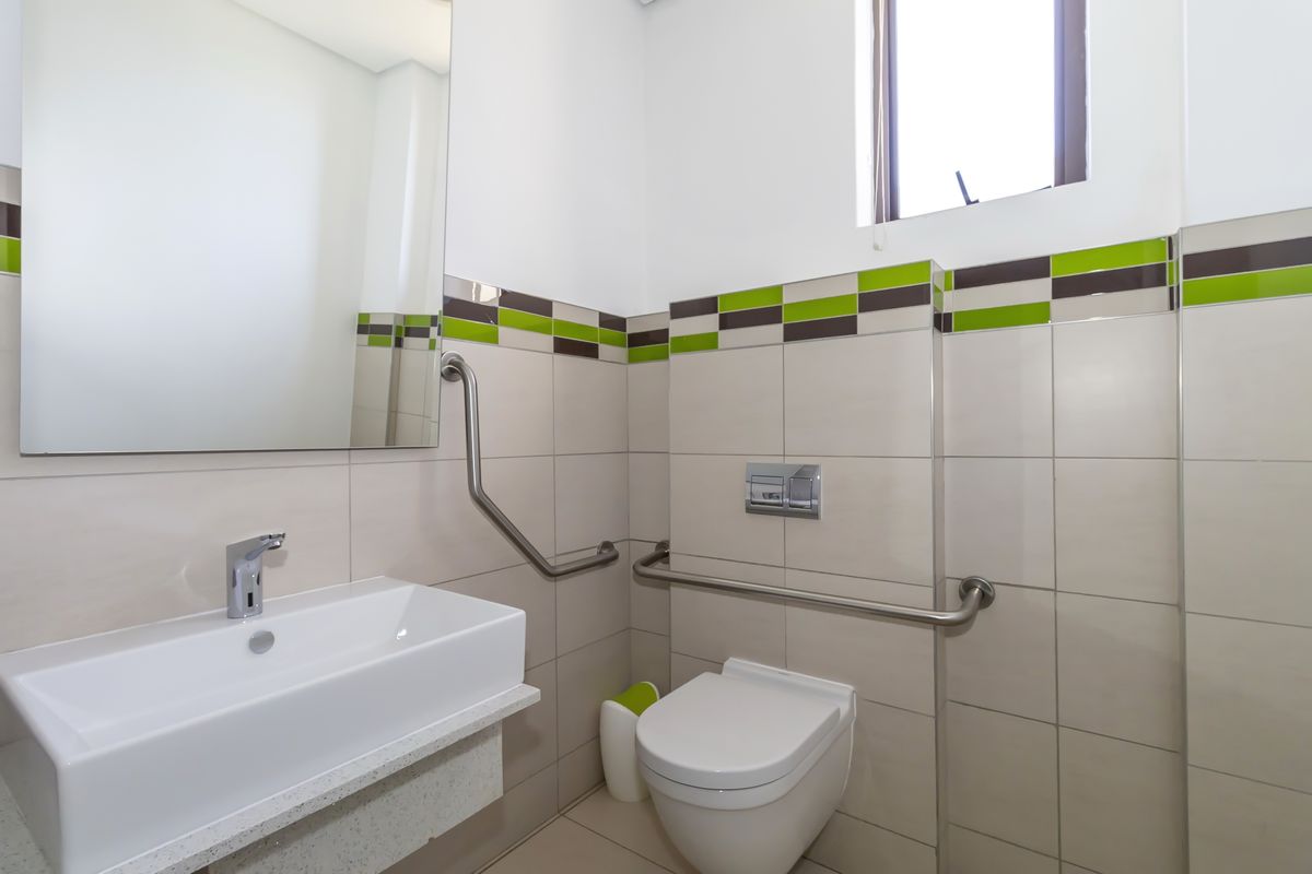 A functional commercial bathroom featuring a wall-mounted toilet, modern basin, and stainless steel grab rails for accessibility, finished with neutral and accent tiling.