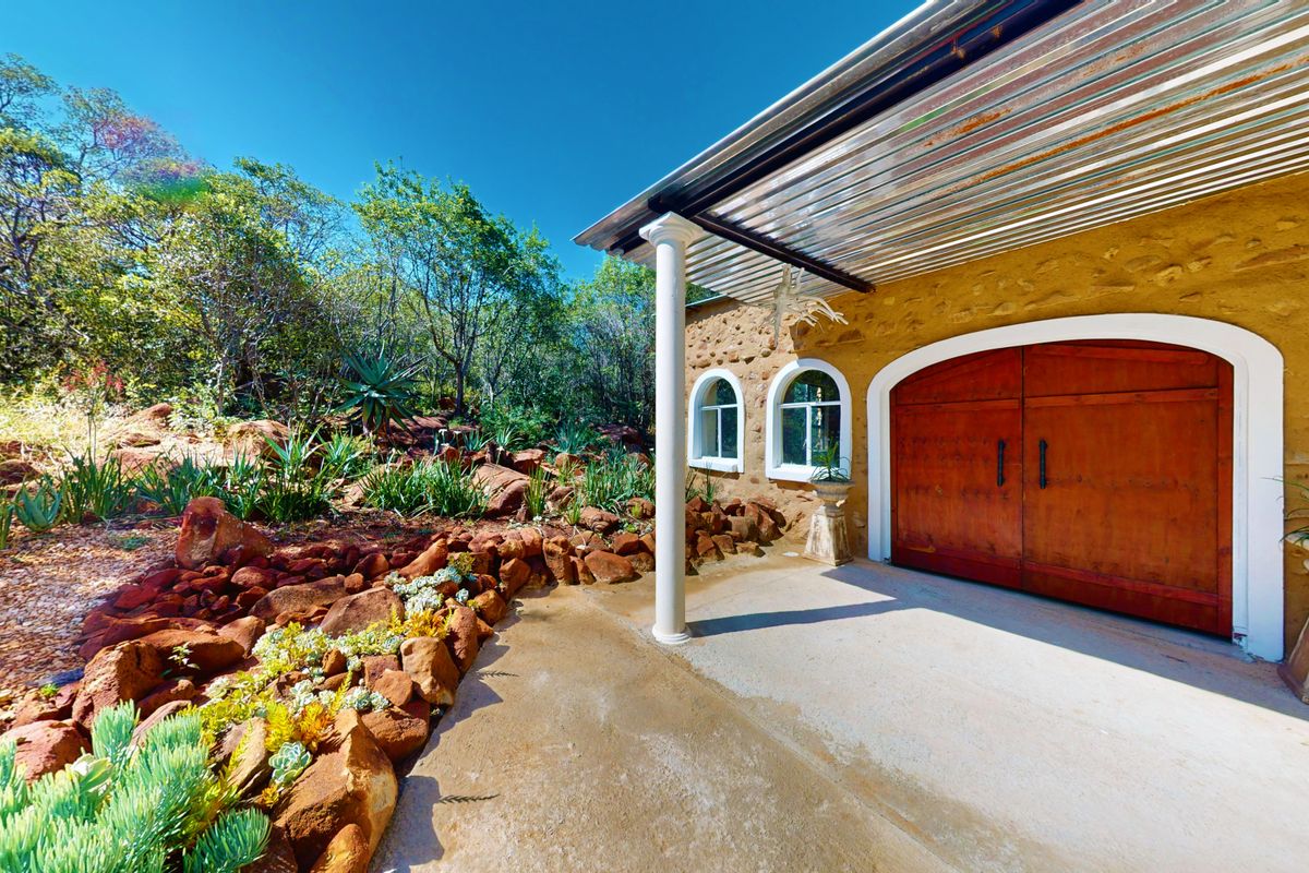 A rustic exterior view featuring a stone facade, arched windows, double wooden doors, and a covered concrete patio area set within a landscaped garden.