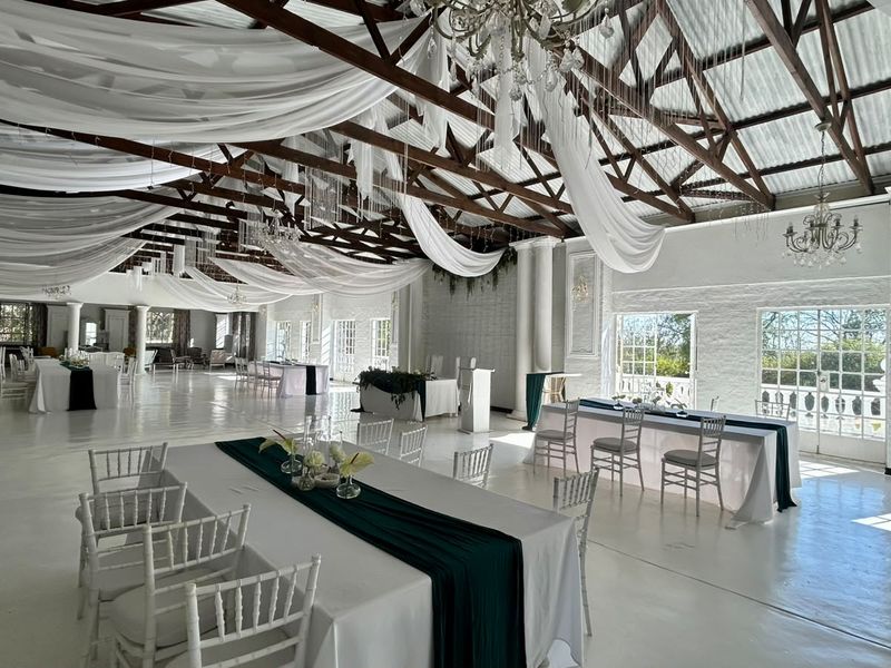 Spacious event venue featuring high vaulted ceilings with exposed timber trusses, elegant chandeliers, painted brick walls, and large windows providing ample natural light.