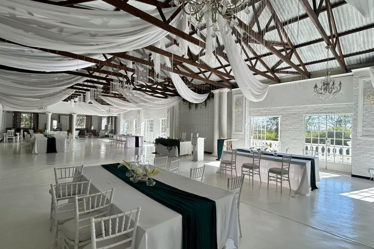 Spacious event venue featuring high vaulted ceilings with exposed timber trusses, elegant chandeliers, painted brick walls, and large windows providing ample natural light.