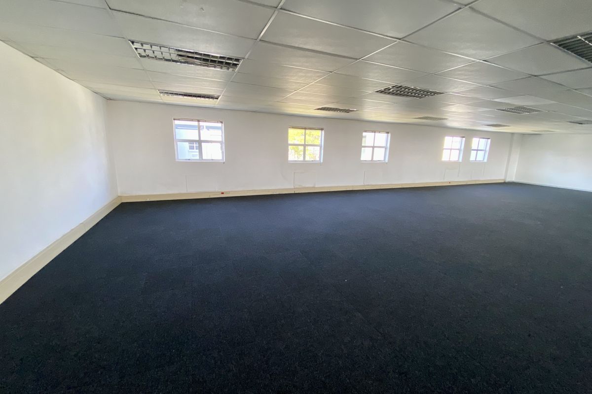 A spacious, open-plan office area featuring a suspended ceiling with integrated lighting, dark carpeting, and multiple windows providing natural light.