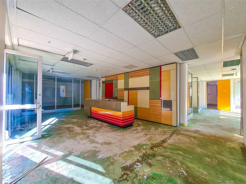 An empty office reception area featuring a built-in desk, glass partitions, and a suspended ceiling, currently requiring significant flooring repairs and refurbishment.