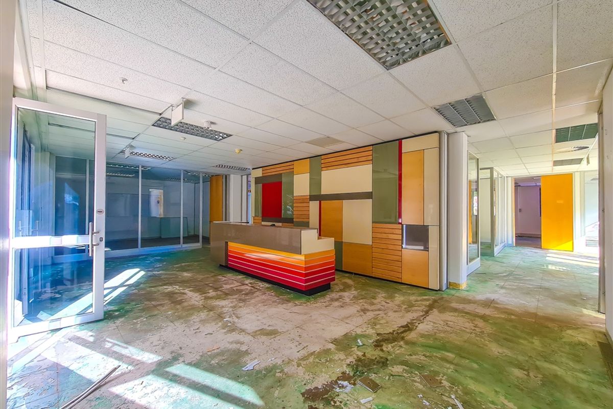 An empty office reception area featuring a built-in desk, glass partitions, and a suspended ceiling, currently requiring significant flooring repairs and refurbishment.