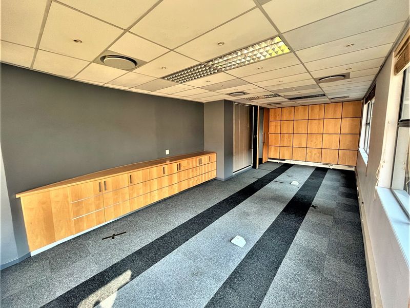 A spacious, empty office featuring built-in wooden cabinetry, wood paneling, carpet tiles, and a suspended ceiling with recessed lighting.