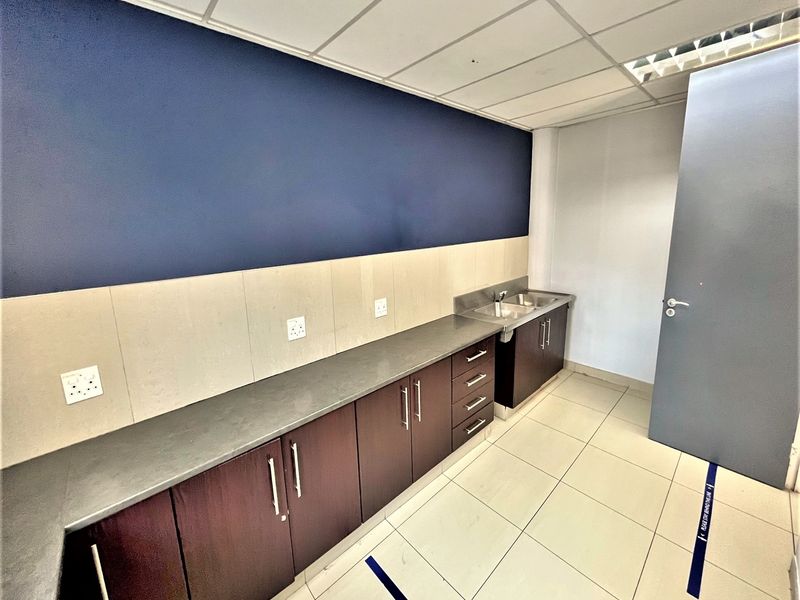 A clean, functional office kitchenette featuring dark wood cabinetry, a stainless steel sink, and neutral tiled flooring under a drop ceiling.