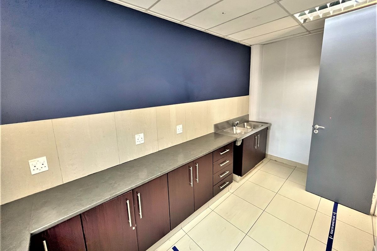A clean, functional office kitchenette featuring dark wood cabinetry, a stainless steel sink, and neutral tiled flooring under a drop ceiling.