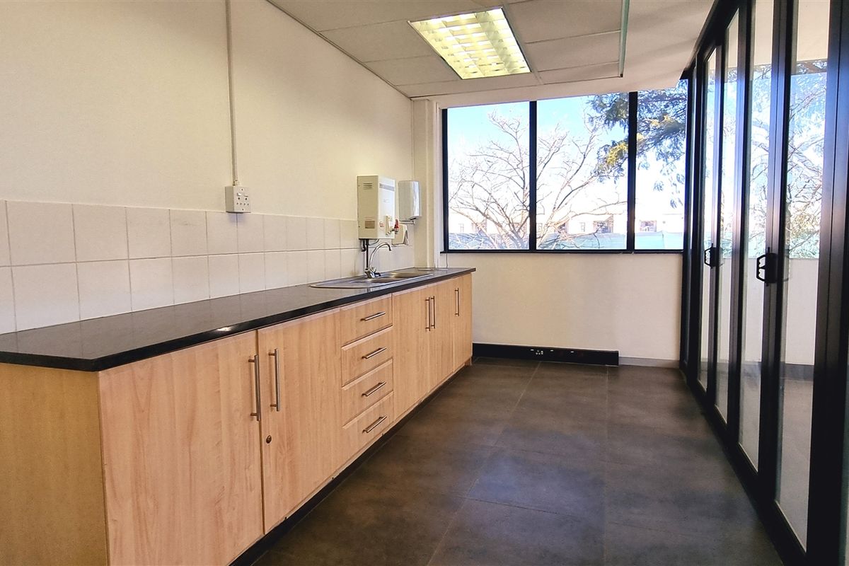 A functional office kitchenette featuring light wood cabinetry, a dark countertop, a stainless steel sink, and large windows providing natural light.