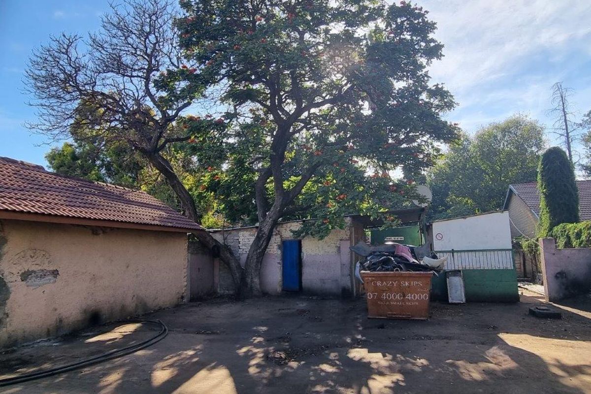 Rear exterior area featuring a paved yard, boundary walls, and a tiled roof structure, showing signs of significant wear and maintenance requirements.