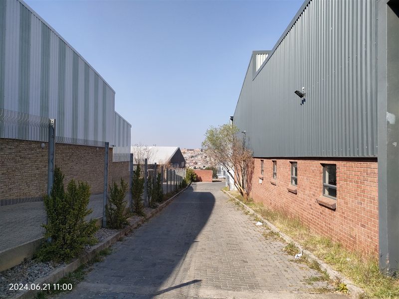 A paved service alleyway between two industrial warehouse buildings featuring brick and corrugated metal construction with perimeter security fencing.