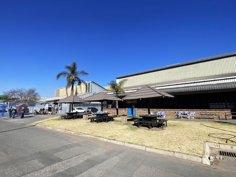 Exterior view of an industrial facility featuring metal cladding, a paved driveway, and a landscaped outdoor area with fixed seating umbrellas.