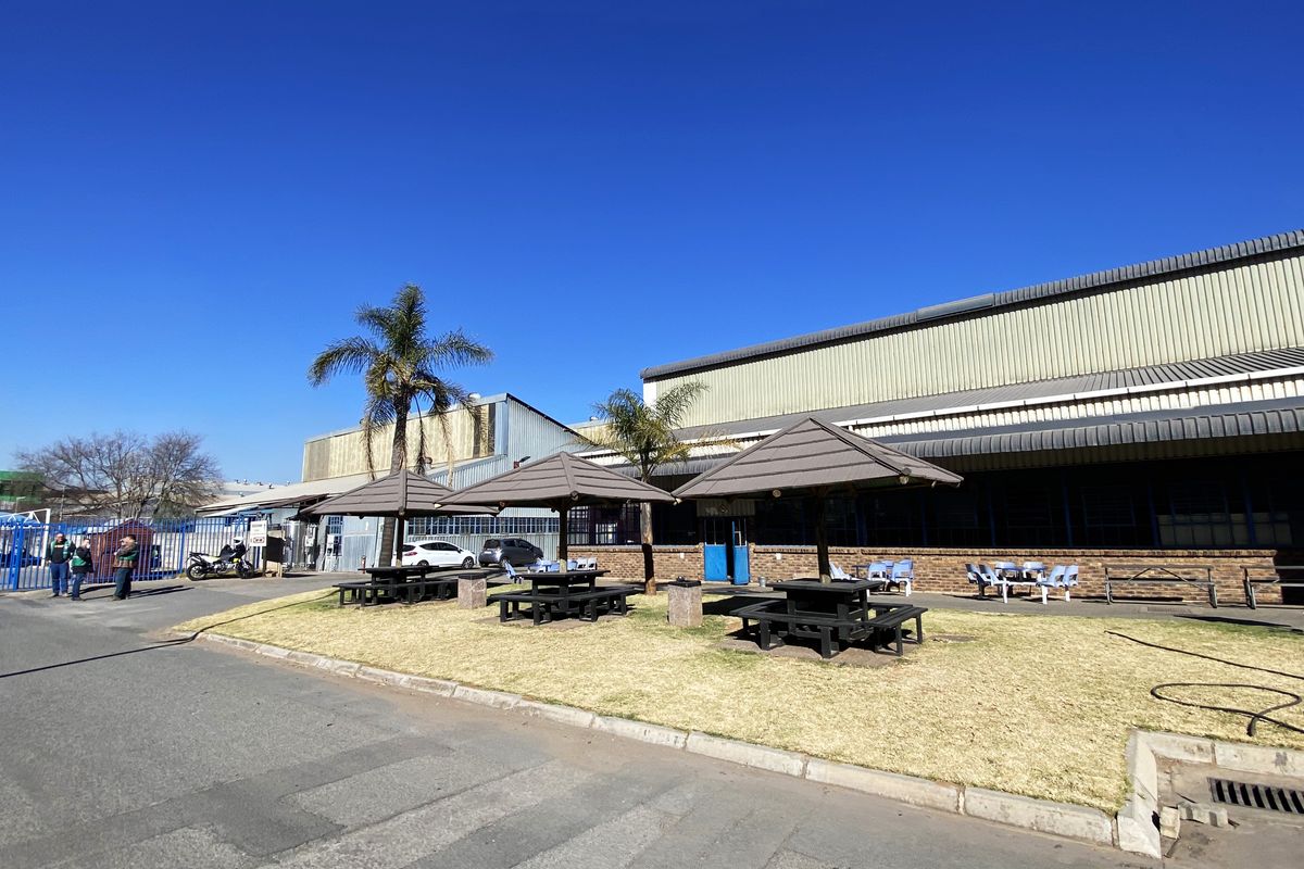 Exterior view of an industrial facility featuring metal cladding, a paved driveway, and a landscaped outdoor area with fixed seating umbrellas.