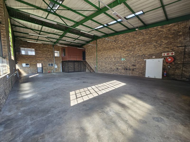 Spacious industrial warehouse featuring exposed brick walls, high steel truss ceilings with skylights, concrete flooring, and an integrated mezzanine level.
