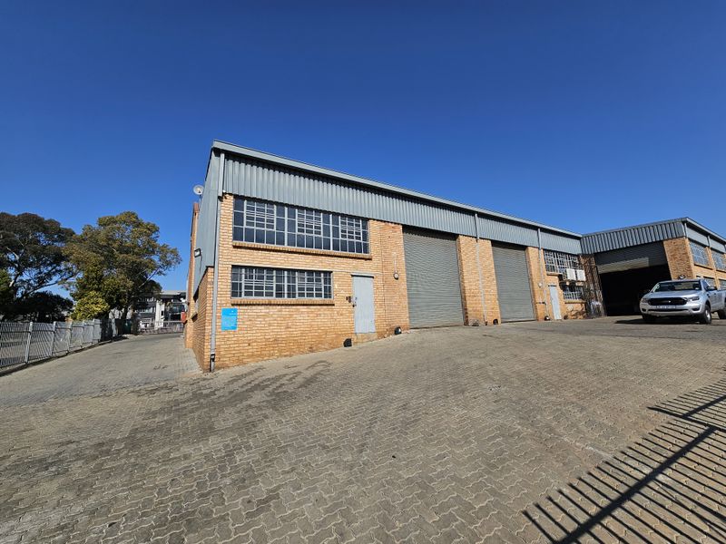 Front exterior view of an industrial warehouse property featuring brick construction, multiple roller shutter doors, and a paved driveway area.