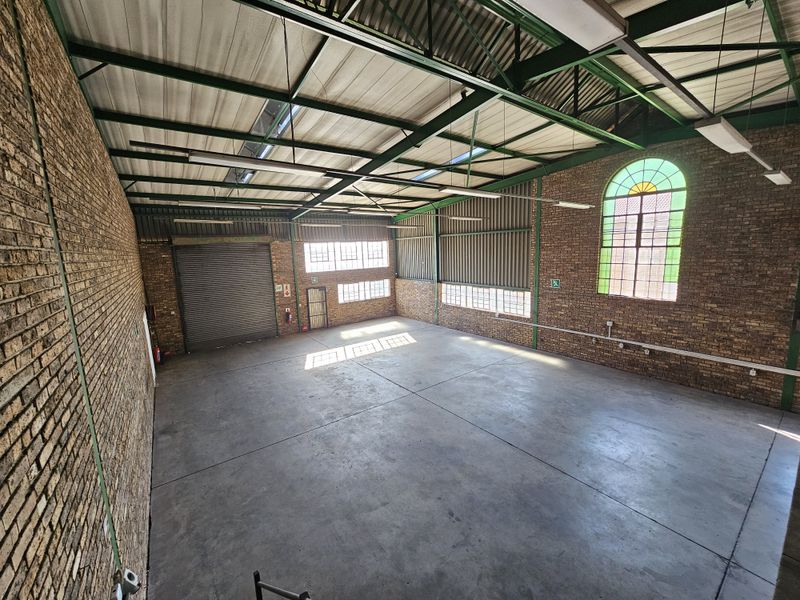 Spacious industrial warehouse unit featuring high exposed steel truss ceilings, concrete flooring, face brick walls, and a large roller shutter door for easy access.