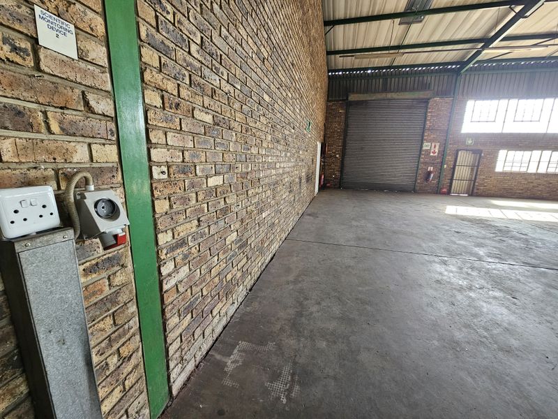 Spacious industrial warehouse unit featuring exposed brick walls, concrete flooring, high ceilings with steel trusses, and a large roller shutter door for easy access.