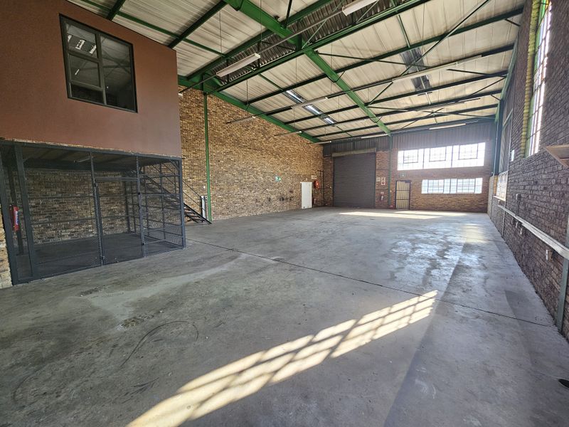 Spacious industrial warehouse unit featuring high ceilings with exposed steel trusses, concrete flooring, a mezzanine level, and a secure roller shutter door.