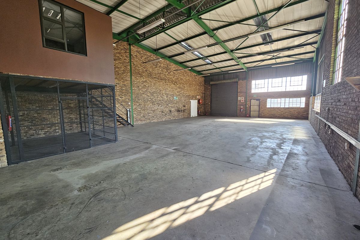 Spacious industrial warehouse unit featuring high ceilings with exposed steel trusses, concrete flooring, a mezzanine level, and a secure roller shutter door.