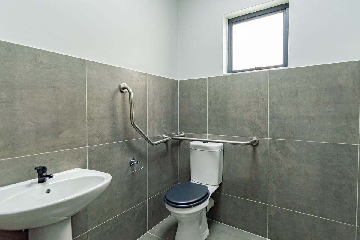 A modern, accessible bathroom featuring large-format grey wall tiles, a pedestal basin, a toilet, and stainless steel grab rails.