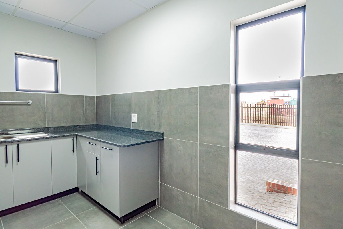 A clean, modern industrial kitchenette featuring grey cabinetry, granite countertops, a stainless steel sink, and large tiled splashbacks with ample natural light.