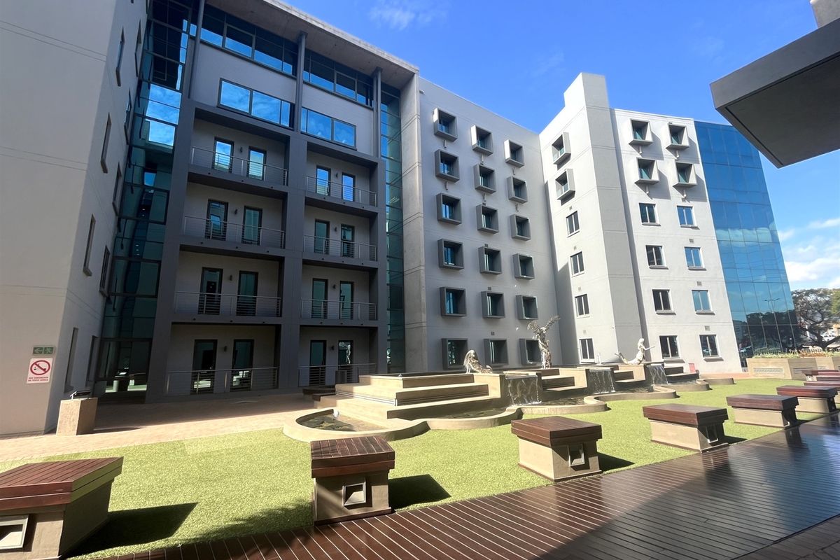 A modern multi-story office building exterior featuring a landscaped courtyard with water features, paved walkways, and integrated seating areas.