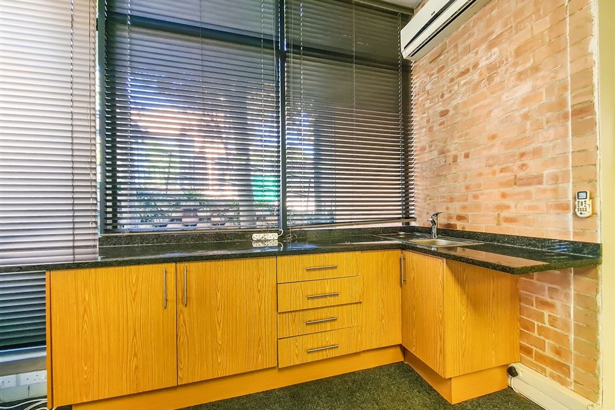 Office kitchenette featuring wood-look cabinetry, granite countertop with integrated sink, exposed brick feature wall, and wall-mounted air conditioning unit.