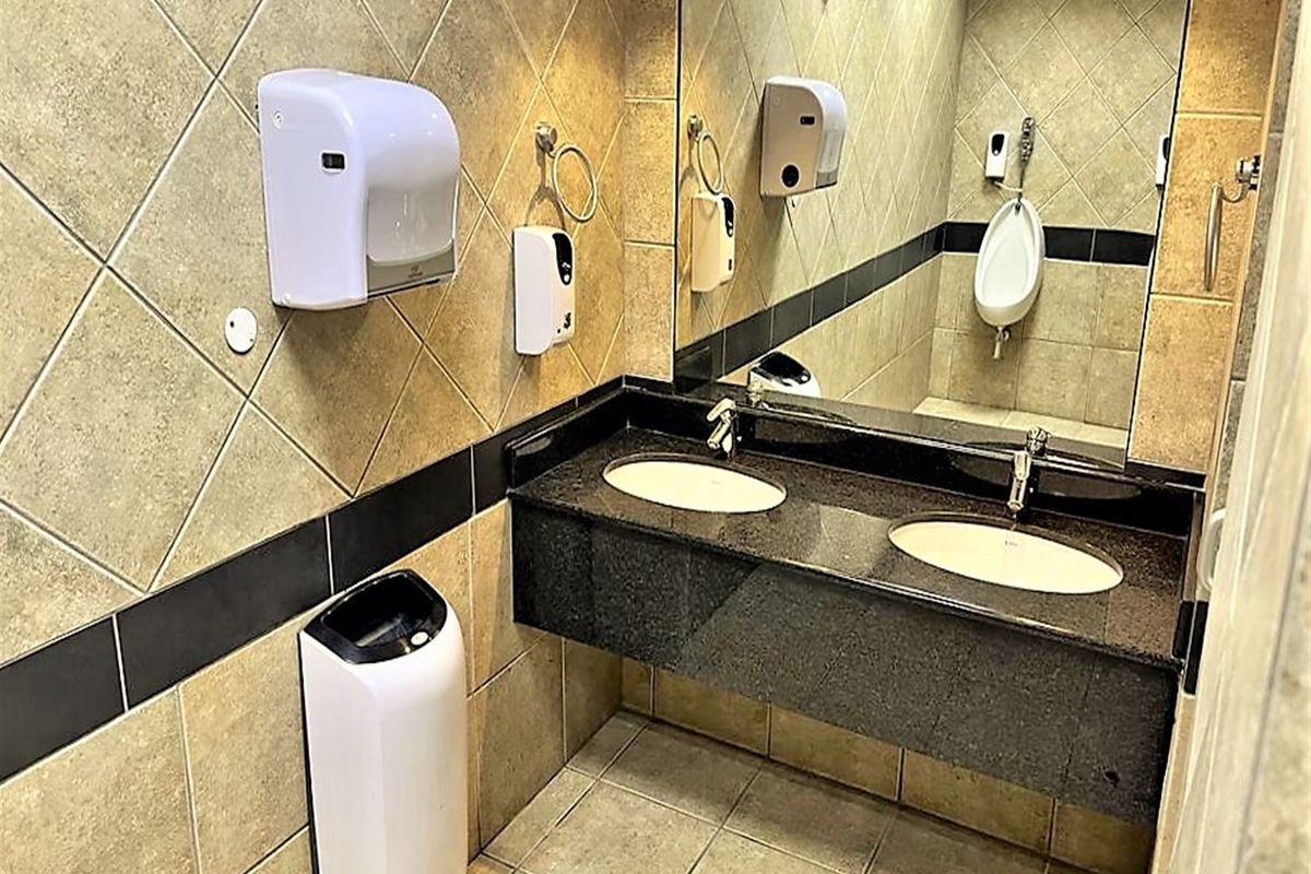 A functional office restroom featuring a double vanity with granite countertop, wall-mounted soap and paper towel dispensers, and tiled wall finishes.