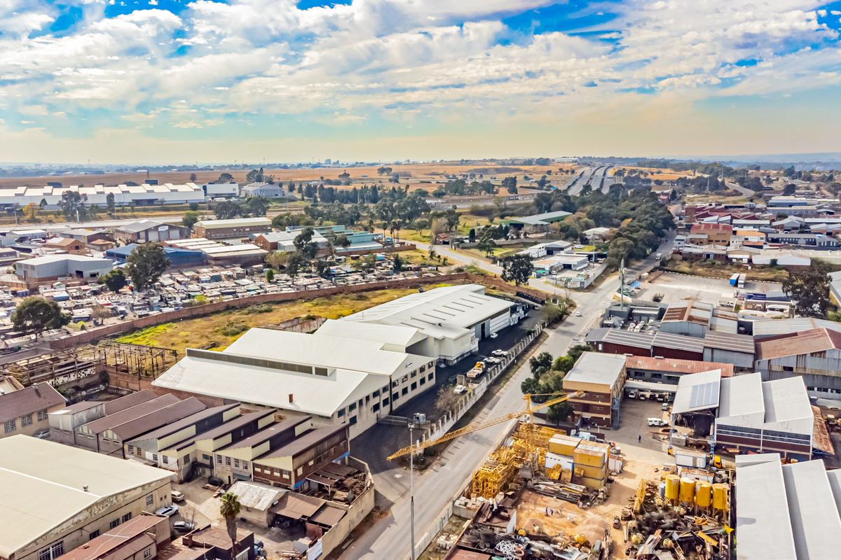 Aerial view of an industrial property complex featuring large warehouse structures with metal roofing, paved yard space, and perimeter fencing in an industrial zone.