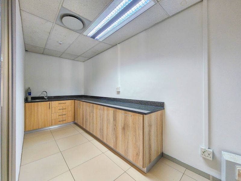 A clean, functional office kitchenette featuring light wood-grain cabinetry, dark granite countertops, a stainless steel sink, and modern drop ceiling lighting.