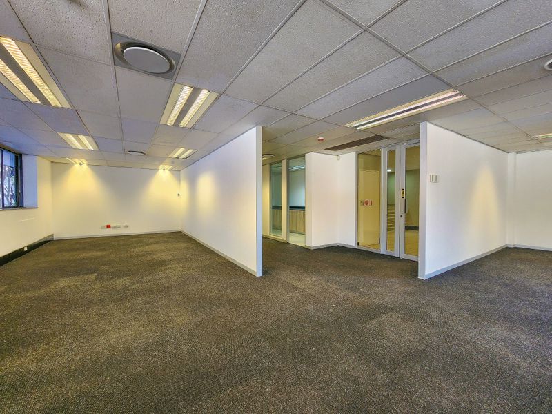 A spacious, vacant office area featuring a suspended ceiling with recessed lighting, carpeted flooring, and glass partition walls creating a professional, open-plan workspace.