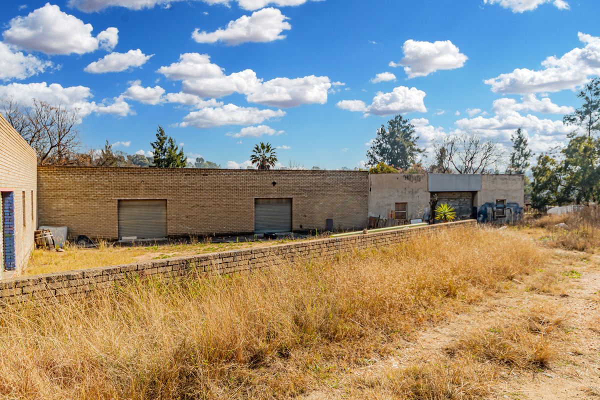 Rear view of a small holding property featuring brick outbuildings with roller shutter doors and an expansive, unmaintained yard area.