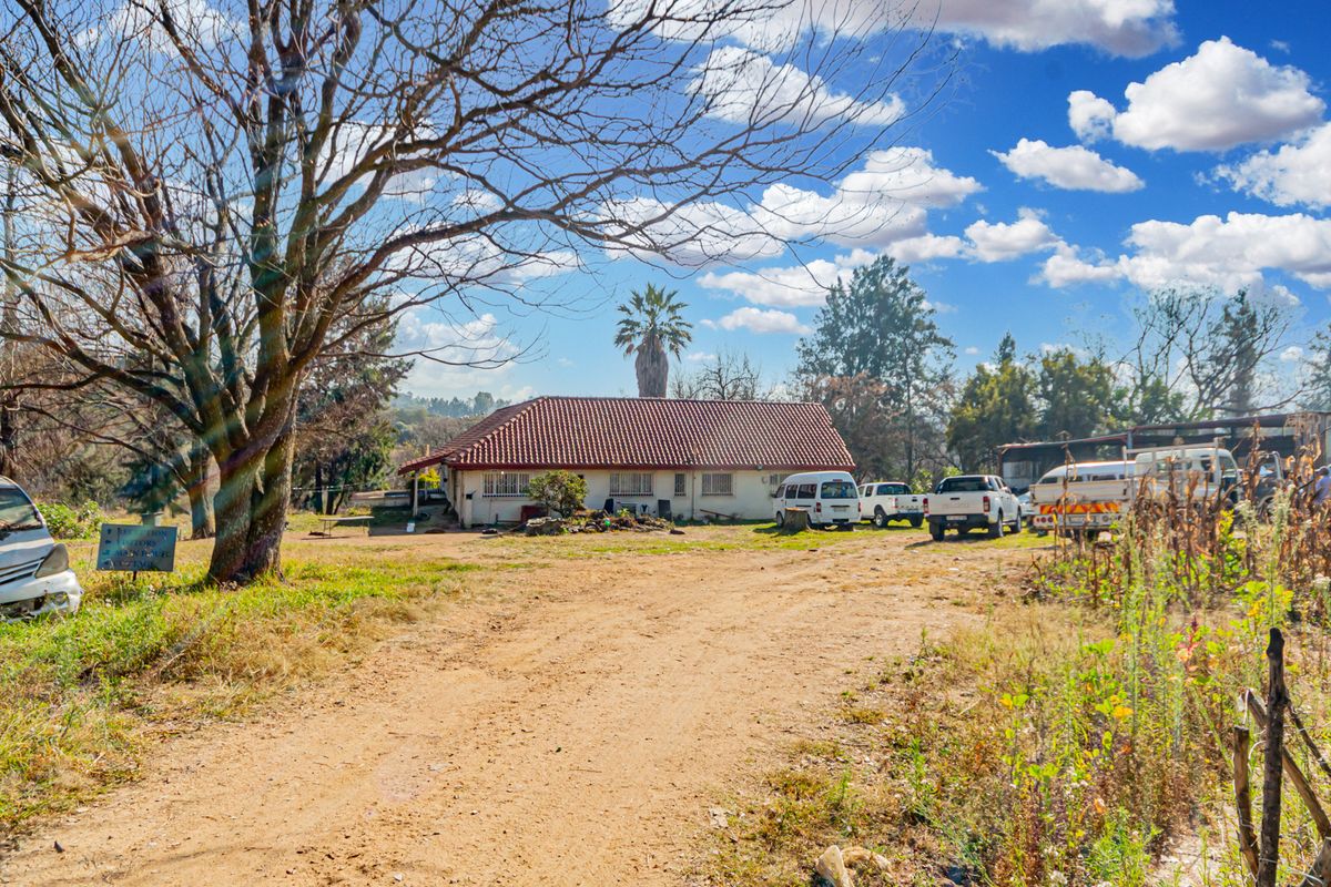 Front view of a small holding property featuring a tiled roof dwelling, security bars, and an unpaved driveway set within a rural landscape.