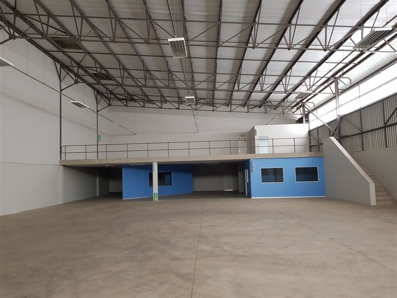 Spacious industrial warehouse featuring a mezzanine level, high steel roof trusses, concrete flooring, and integrated office or storage rooms with blue painted walls.